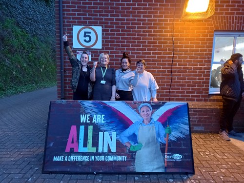 Four women from Colchester domestic abuse charity Next Chapter are stood behind an All In banner. They are celebrating receiving a £750 grant.