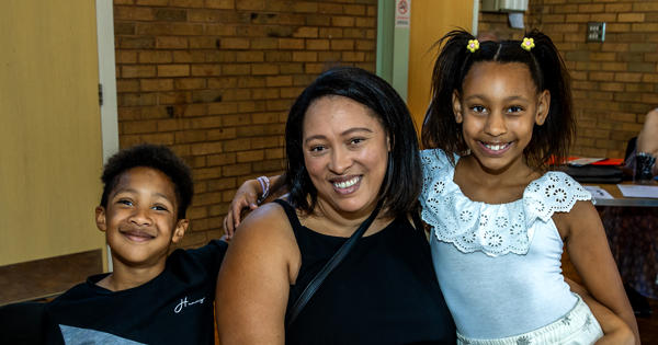 A woman is sat at a table. Her two children, a young boy to her left and young girl to her right, are stood next to her.