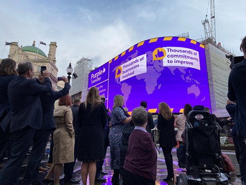 A group of Purple Tuesday partners standing and facing the Piccadilly Circus Lights billboard display, which is lit up in purple and says "Thousands of commitments to improve"