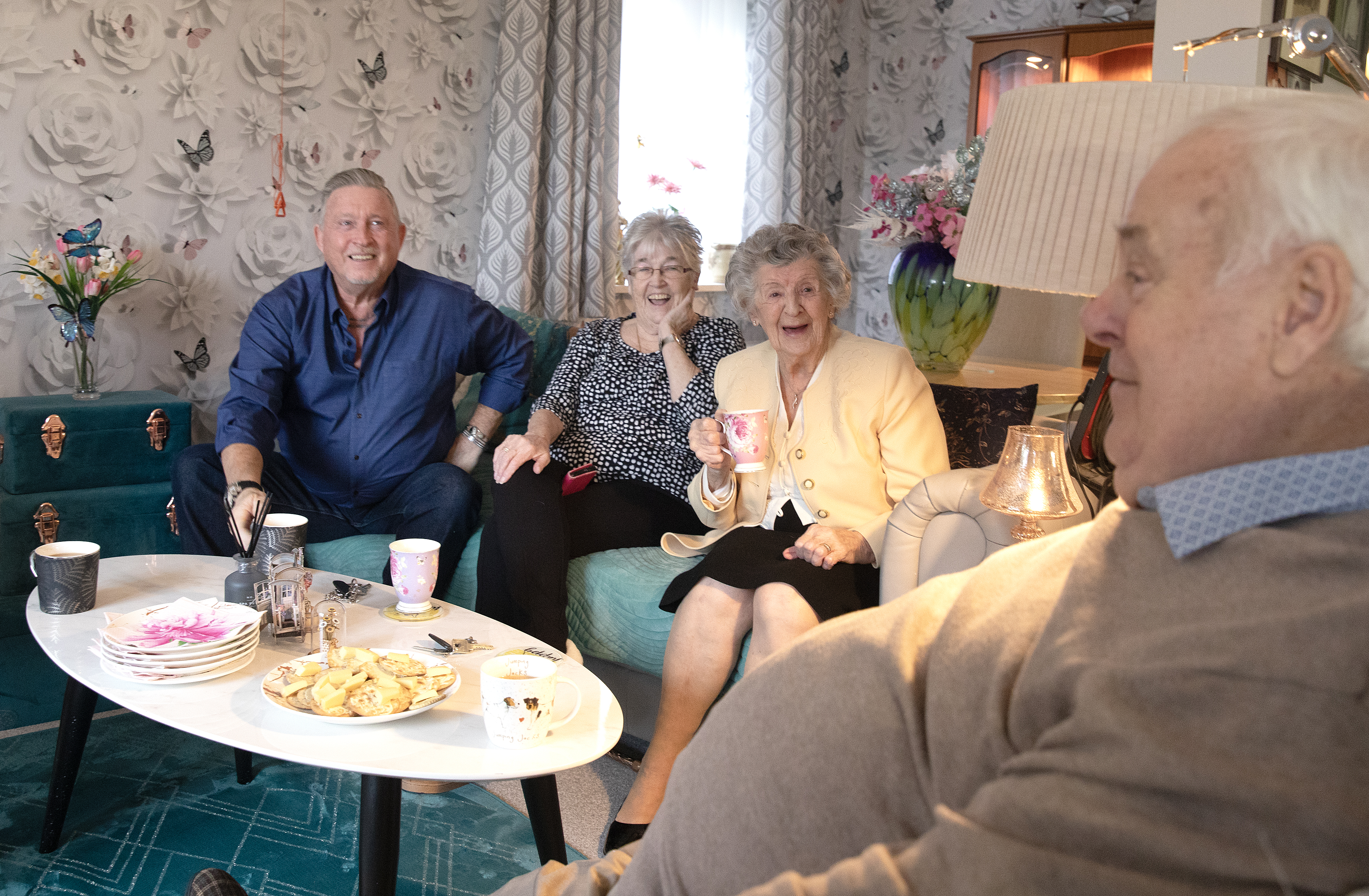 Eastlight resident Martin (on the left-hand side of the photo), sitting together with a group of residents inside the living room of one of our housing for older people schemes. The four residents are sitting on chairs around a small table, which has tea and cake on it. They are smiling and appear to be enjoying a joke together.