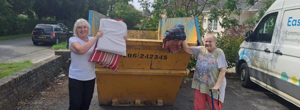 Two Eastlight residents standing in front of a skip, proudly holding up items about to be thrown into it. To the right of the skip is an Eastlight van. The residents were photographed during a clean-up event on their local estate. In the background is a road, some houses and a car parked on the street. 