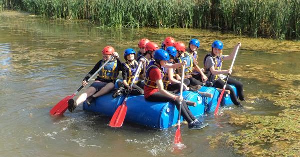 Young people from Braintree Youth Project Charity sat on a handmade craft floating on a river. They are all wearing life jackets and helmets and holding paddles to move them along the river.