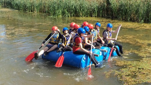 Young people from Braintree Youth Project Charity sat on a handmade craft floating on a river. They are all wearing life jackets and helmets and holding paddles to move them along the river.