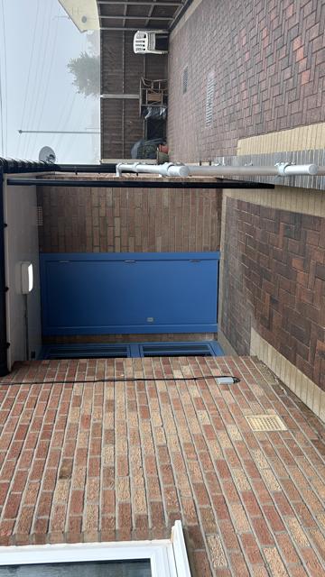 A newly paved ramp leading to a blue door. The ramp is wide and a wall has been built next to the ramp with silver railings on top.