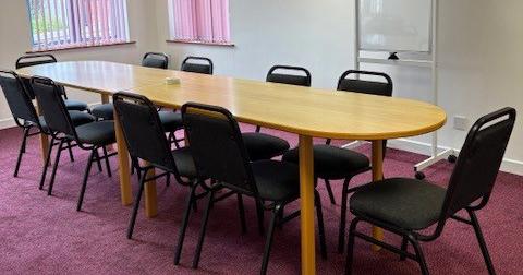A large room features a long oval desk with 10 chairs around it. There is also a freestanding white board and two sets of windows letting in light.