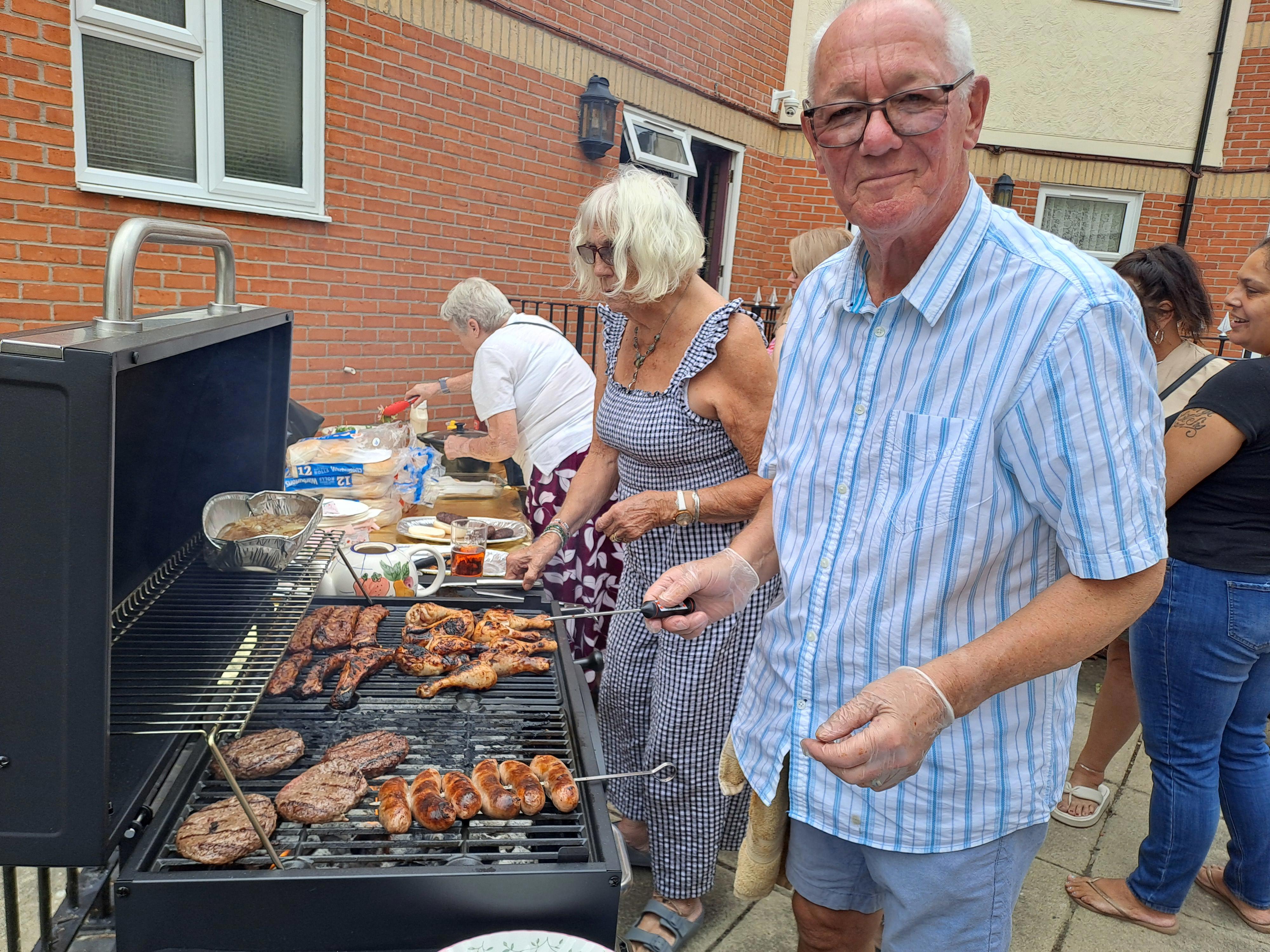 An older man and woman are stood next to a barbeque with barbeque tools in their hand. The man is looking at the camera and smiling.
