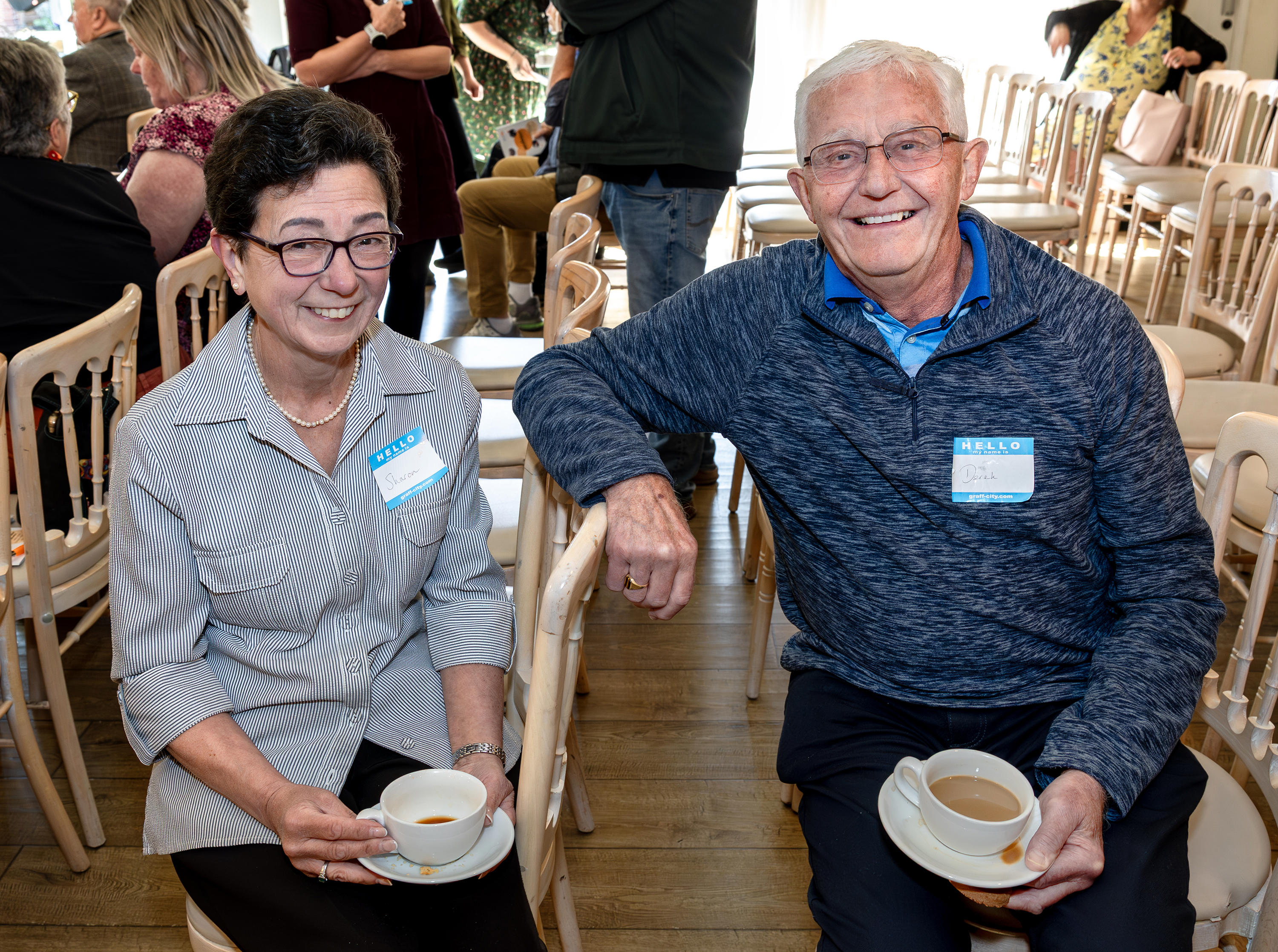 A woman (on the left) and a man (on the right) are sat next to each other. They are smiling and are each holding a cup of tea and saucer.