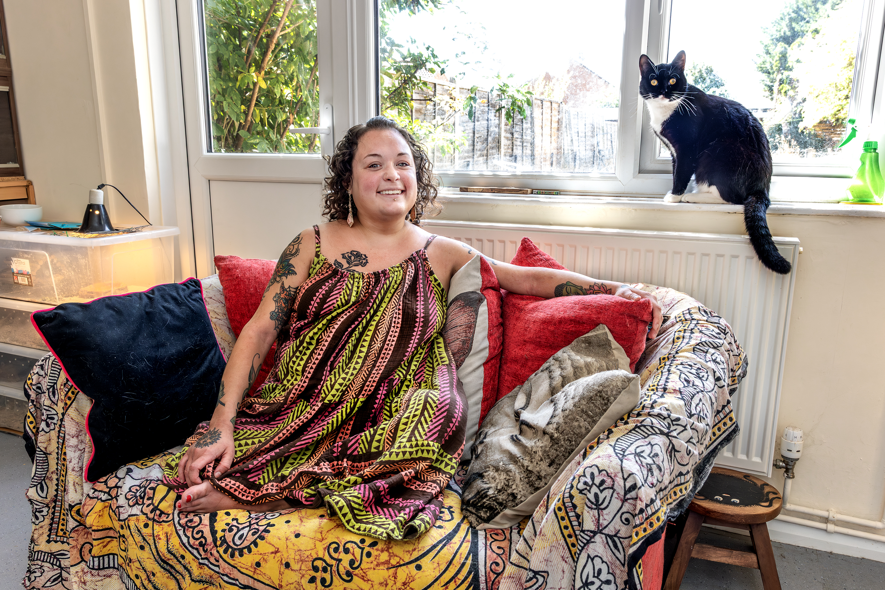 Eastlight resident, Bobbie, is sat on a sofa in her home. Her black and white cat is sat on the window sill behind her. They are both looking at the camera.