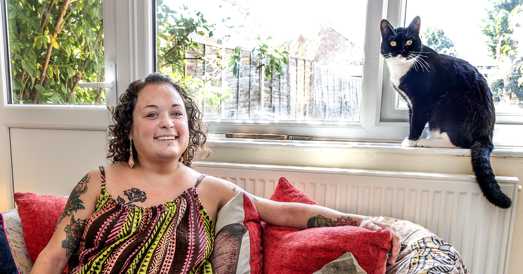 Eastlight resident, Bobbie, is sat on a sofa in her home. Her black and white cat is sat on the window sill behind her. They are both looking at the camera.