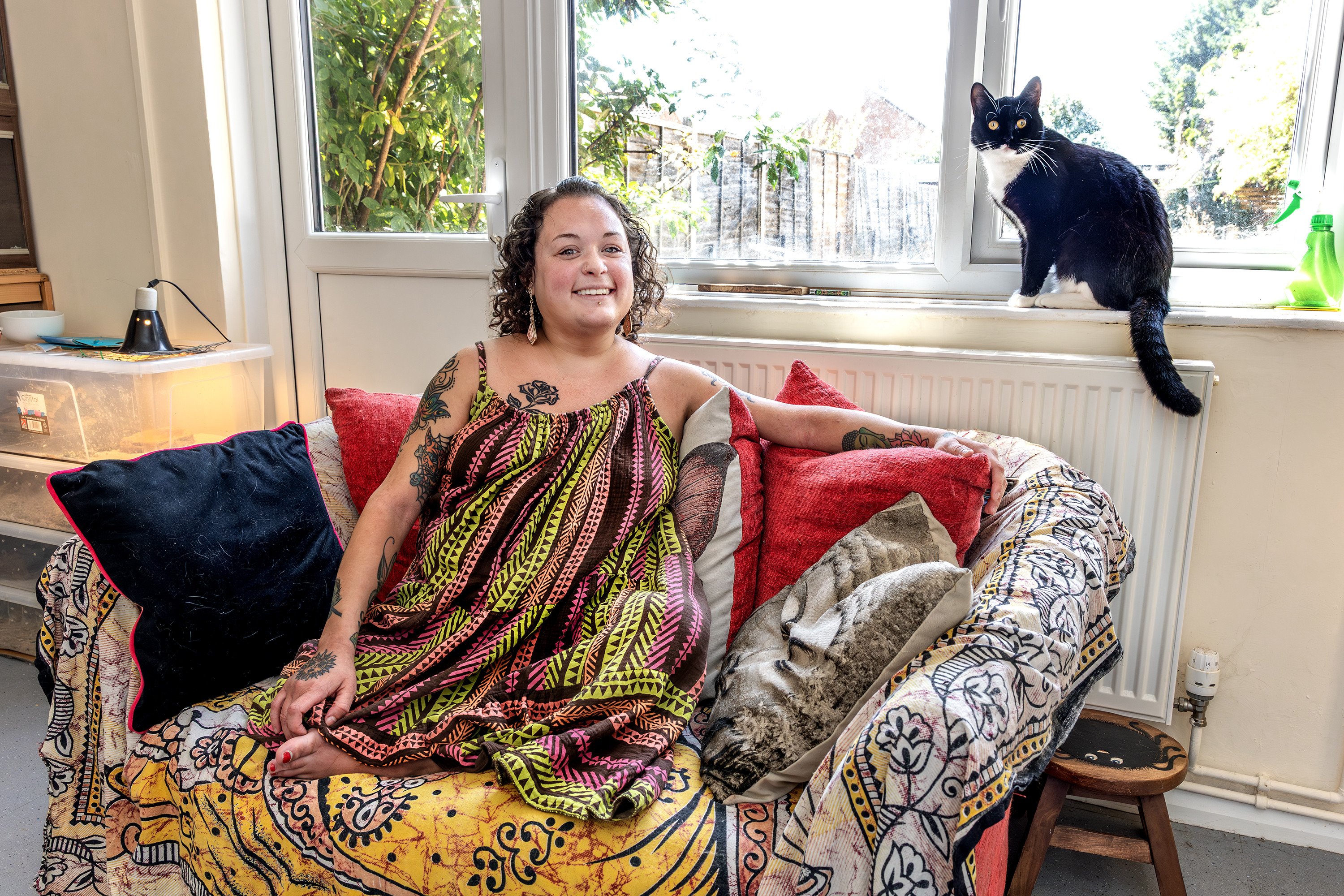 Eastlight resident, Bobbie, is sat on a sofa in her home. Her black and white cat is sat on the window sill behind her. They are both looking at the camera.