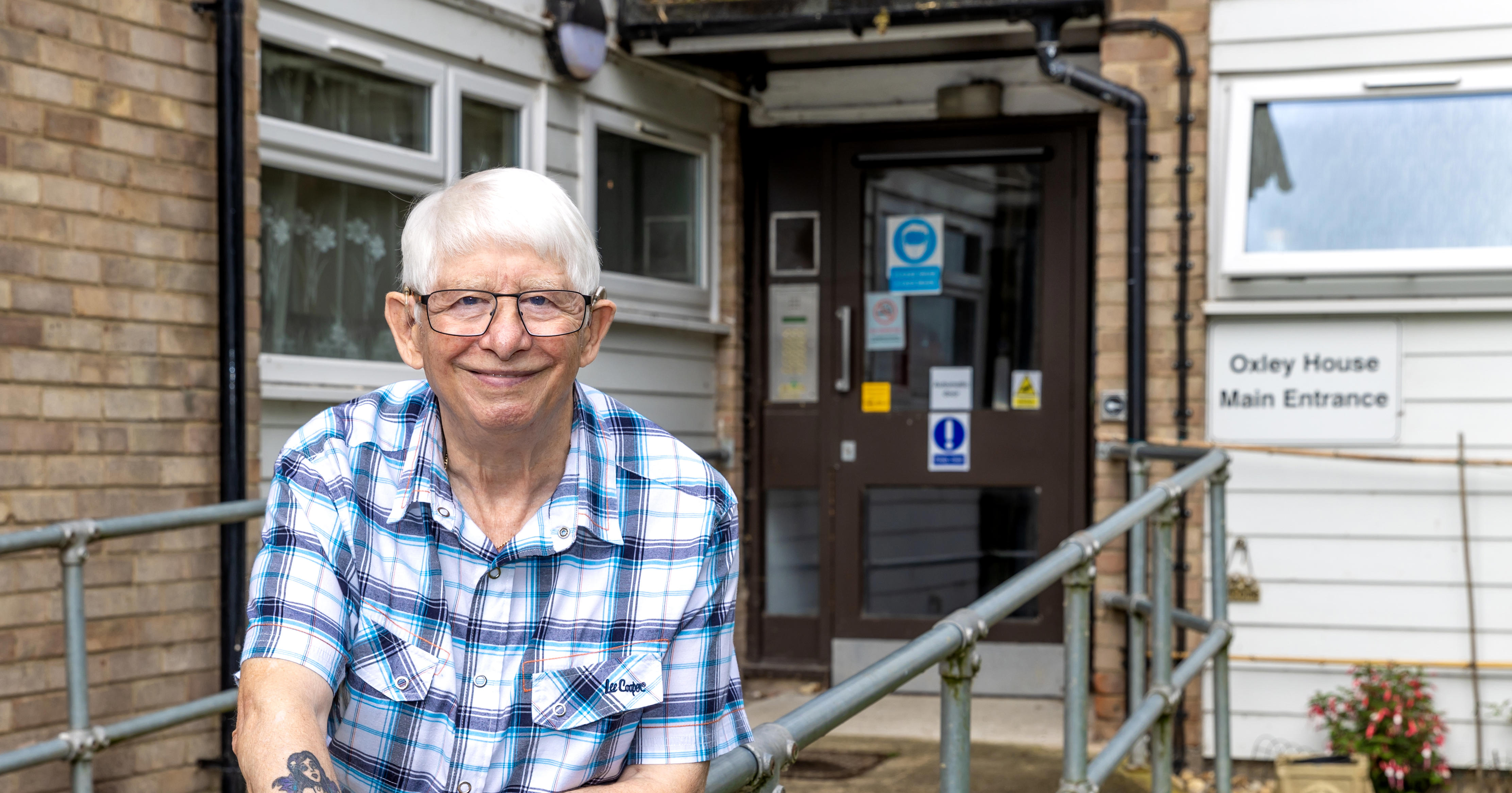 An older man with glasses is leaning on a metal railing which leads up a ramp to a block of flats.