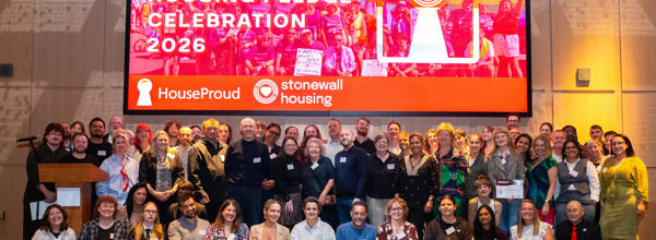 A group photo of award winners and attendees at the LGBTQ+ Housing Pledge Celebration Awards 2026, standing in front of the event stage and screen.