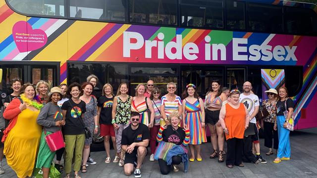 Members of Black Cactus Choir are standing in front of a Pride In Essex bus at Colchester Pride 2023. All 23 members are dressed colourfully and smiling at the camera.