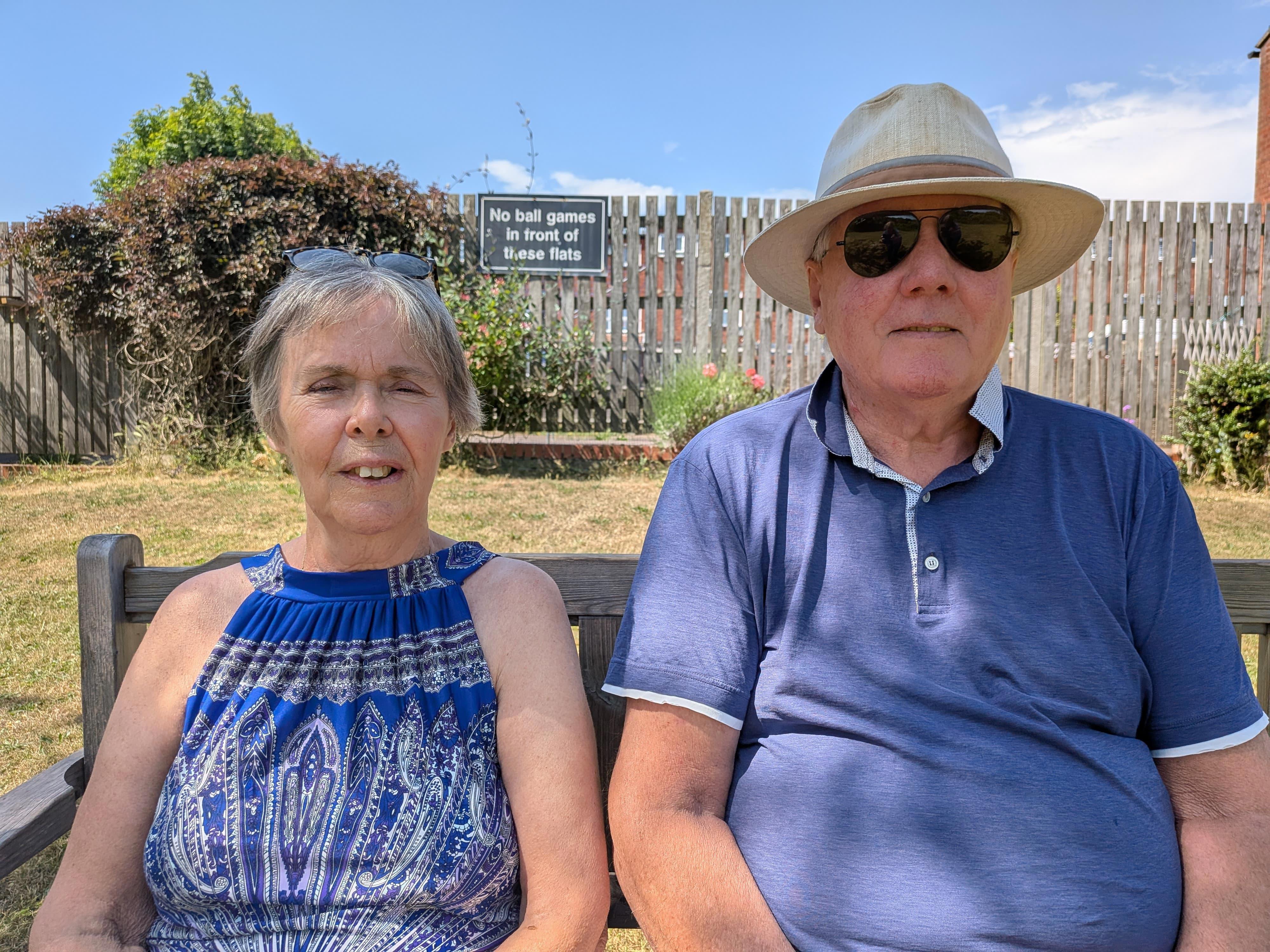 Tim and Liz Golden sitting on a bench in their communal garden. They are wearing blue tops, and Tim wears a Panama-style hat. There is grass and a wooden fence in the background, set beneath a bright blue sky. 