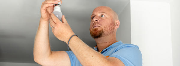 A member of Eastlight's Home Solutions Team replaces a ceiling light bulb during a home safety visit. The image shows a bright, modern room with a smoke alarm visible on the ceiling.