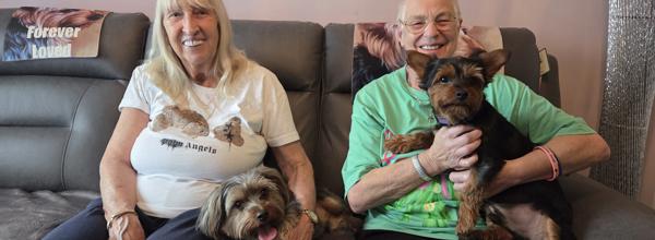 Two older female Eastlight residents sitting together on a sofa in a living room, smiling at the camera. Each resident is holding a small, terrier-type dog on their laps. 