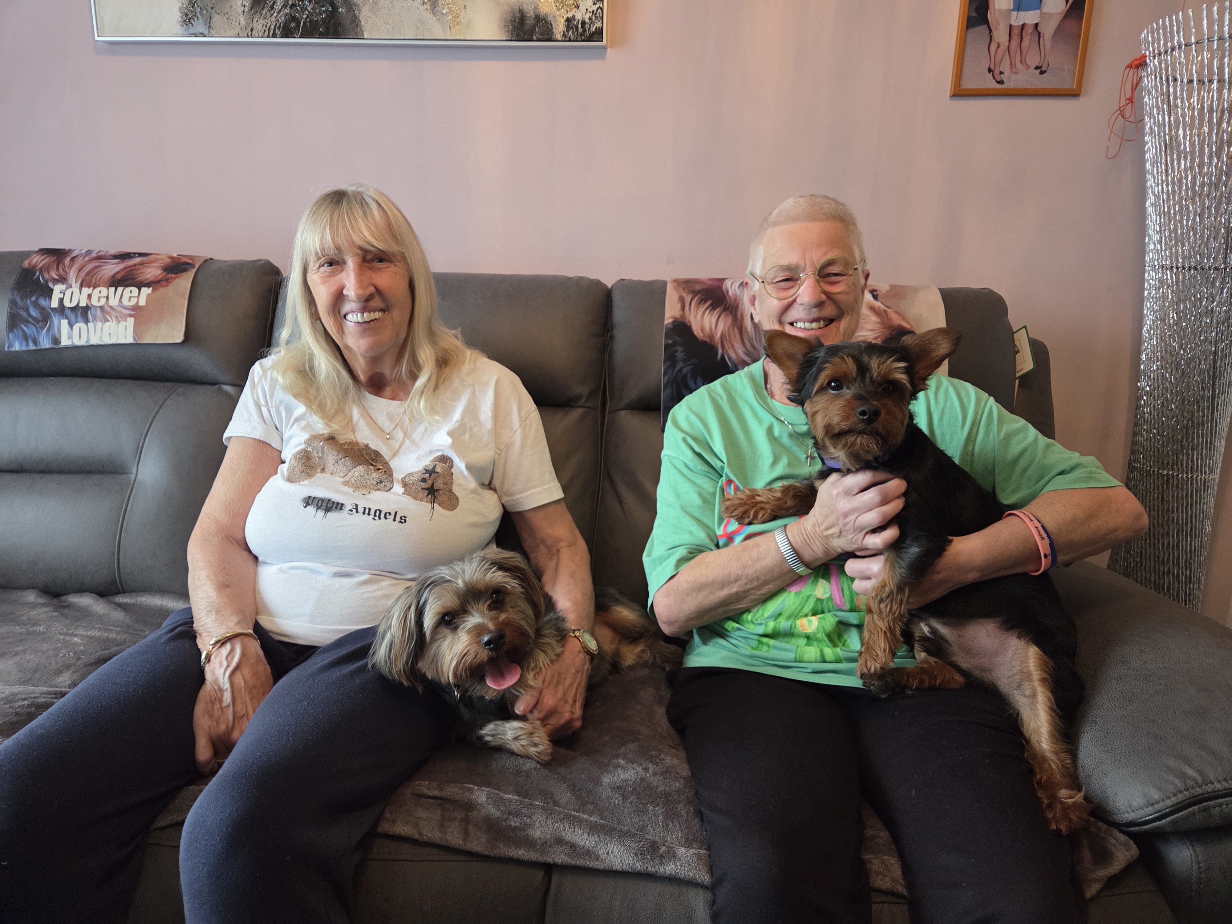 Two older female Eastlight residents sitting together on a sofa in a living room, smiling at the camera. Each resident is holding a small, terrier-type dog on their laps. 