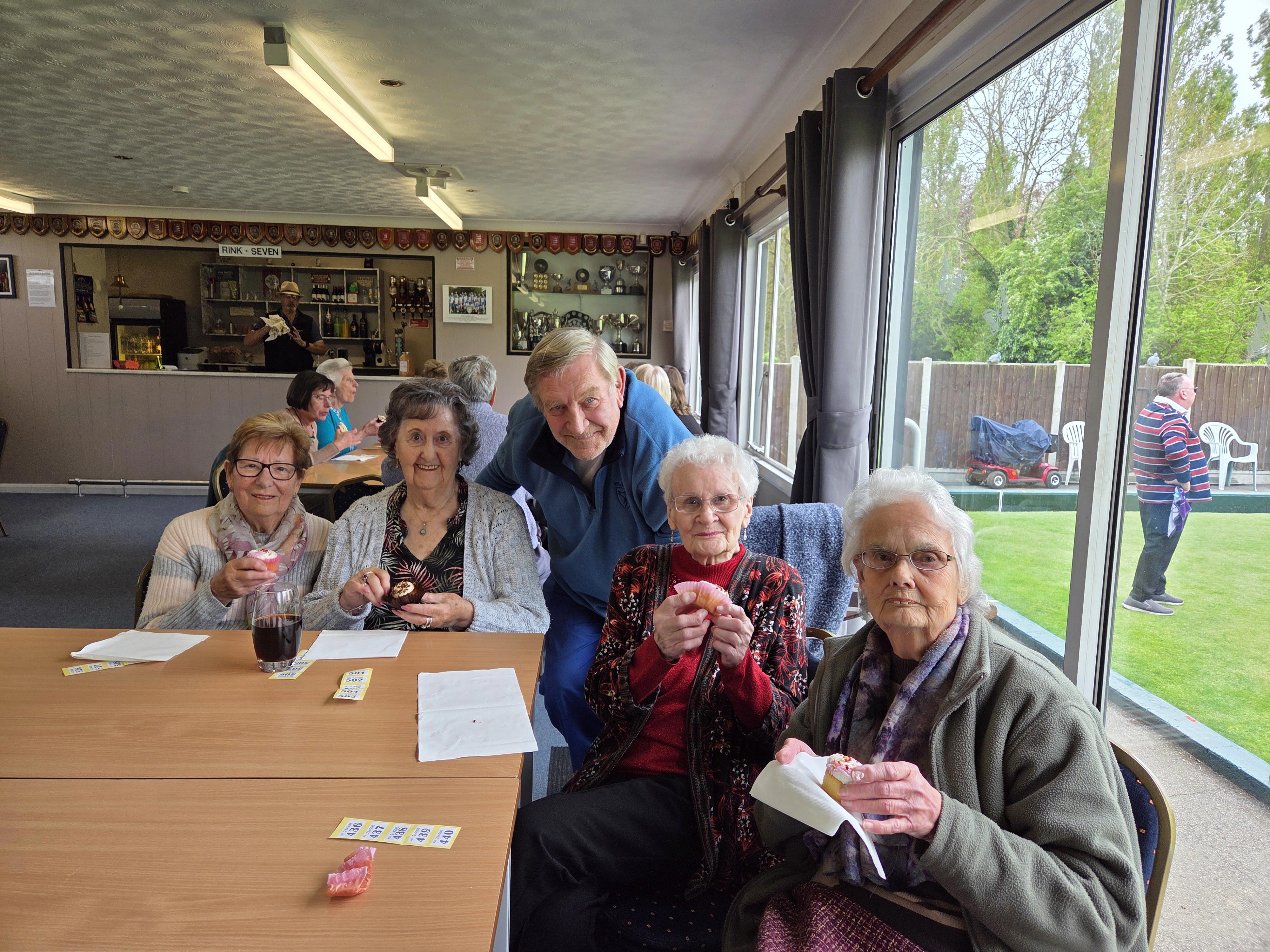 A group of four older ladies and an older man are sat together. The older women are each holding a cup of tea.
