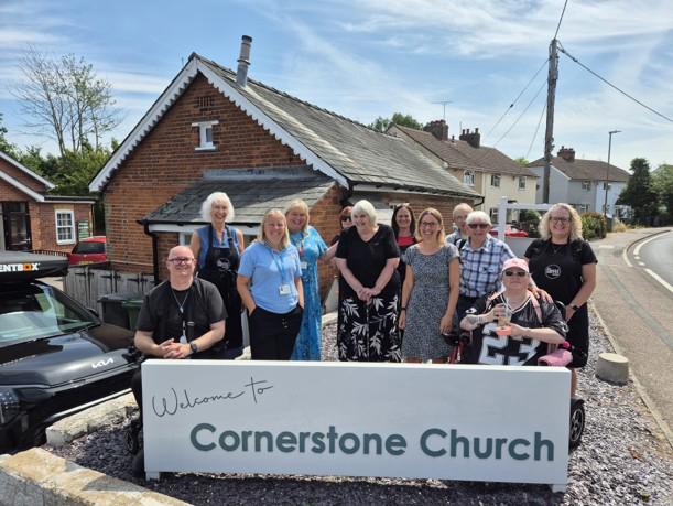 A group of Eastlight residents stood together on the street, smiling at the camera, behind a low-hanging sign which says 'Welcome to Cornerstone Church.'  In the background are some houses dotted along what appears to be a main road. 