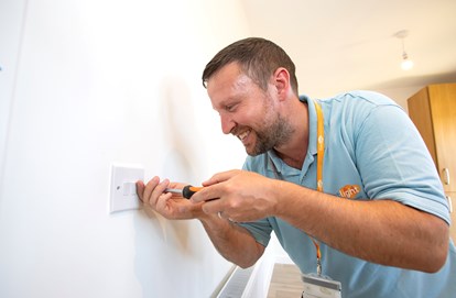 An electrician is unscrewing a light switch. He is wearing an Eastlight polo shirt and lanyard.