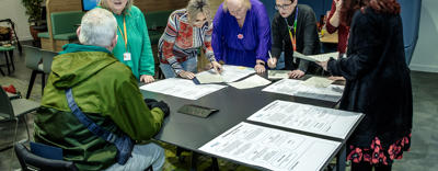 A small group of residents and Eastlight staff (seven people in total) are stood gathered around a table/office desk inside Eastlight House, Braintree. They are chatting and looking down at some large (possibly A3-sized) pieces of paper on the table. The paper contains text (not decipherable in the photo).