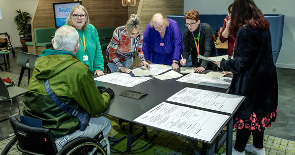 A small group of residents and Eastlight staff (seven people in total) are stood gathered around a table/office desk inside Eastlight House, Braintree. They are chatting and looking down at some large (possibly A3-sized) pieces of paper on the table. The paper contains text (not decipherable in the photo).