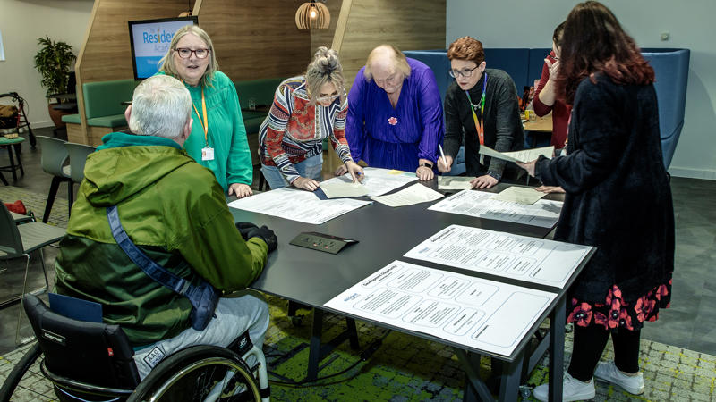A small group of residents and Eastlight staff (seven people in total) are stood gathered around a table/office desk inside Eastlight House, Braintree. They are chatting and looking down at some large (possibly A3-sized) pieces of paper on the table. The paper contains text (not decipherable in the photo).