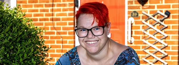 A woman with short red hair and glasses is leaning on a wooden gate in her front garden.