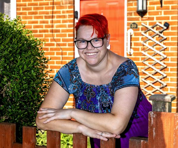 A woman with short red hair and glasses is leaning on a wooden gate in her front garden.