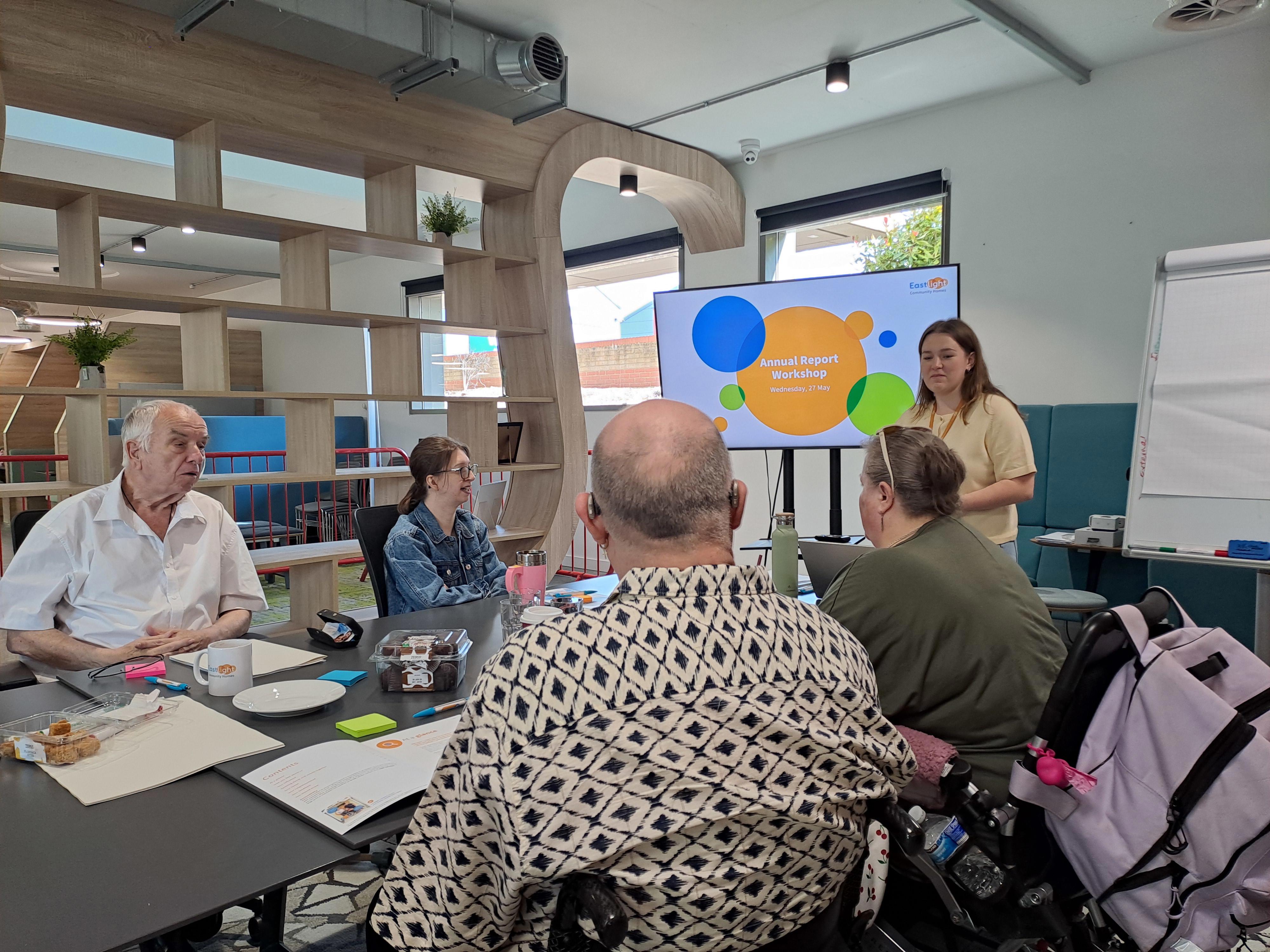 A group of Eastlight residents sitting around a table at Eastlight Hub in Braintree, facing a member of Eastlight staff, who is standing in front of a large screen, smiling at the workshop participants. The screen is showing an Eastlight logo and the words “Annual Report workshop”. To the right of the staff member and the screen is a flipchart with paper. 