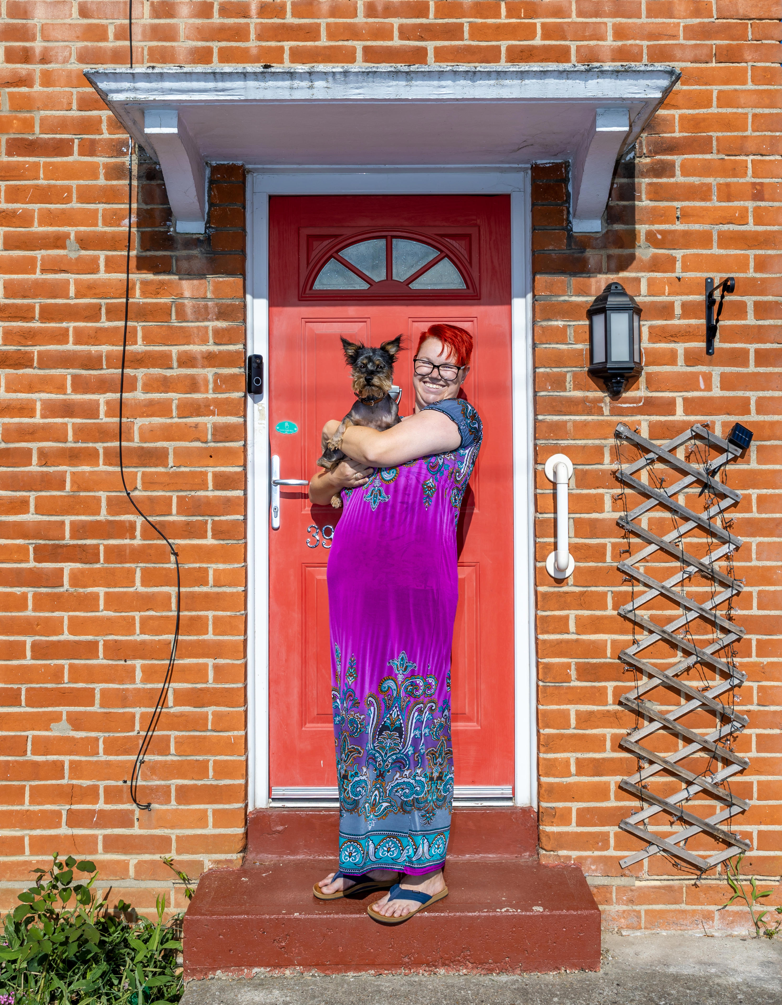 A woman is stood on her doorstep in front of a red front door. She is holding a small yorkshire terrier in her arms.
