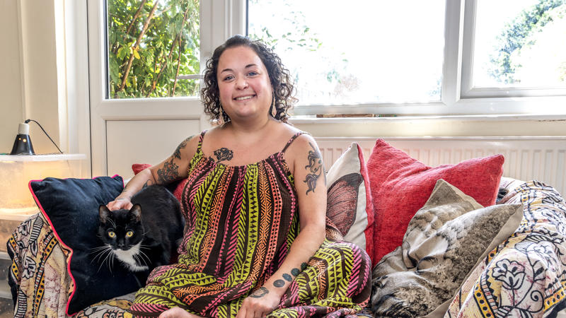 Young resident Bobbie Town is sat on a sofa in her home. Her black and white cat is sat next to her.