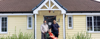 A family consisting of a husband, wife and adult son, pictured on their front doorstep, smiling at the camera. The wife and mother of the family is sitting in a wheelchair, with her husband and son standing behind her, smiling at the camera. 