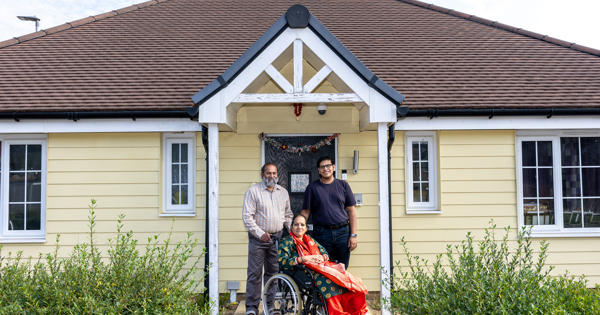 A family consisting of a husband, wife and adult son, pictured on their front doorstep, smiling at the camera. The wife and mother of the family is sitting in a wheelchair, with her husband and son standing behind her, smiling at the camera. 