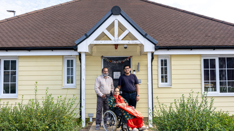 A family consisting of a husband, wife and adult son, pictured on their front doorstep, smiling at the camera. The wife and mother of the family is sitting in a wheelchair, with her husband and son standing behind her, smiling at the camera. 