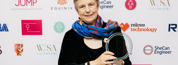 Eastlight's Chair of the Board, Hattie Llewelyn-Davies, holding an award she has won. In the background is a wall covered with various award event sponsor logos. The award held by Hattie is a glass plaque. 