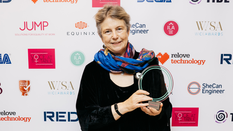 Eastlight's Chair of the Board, Hattie Llewelyn-Davies, holding an award she has won. In the background is a wall covered with various award event sponsor logos. The award held by Hattie is a glass plaque. 