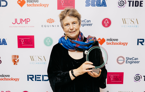 Hattie standing in front of a branded conference banner (with lots of partner logos on it). She is holding her award, smiling at the camera. She is wearing a black dress and a colourful scarf.