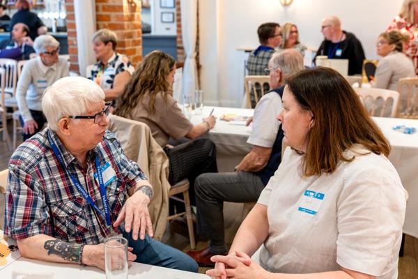 A  resident talking with a staff member during the Eastlight Resident Members Charter celebration. In the background, there are more tables and chairs, with other staff members and residents chatting. 