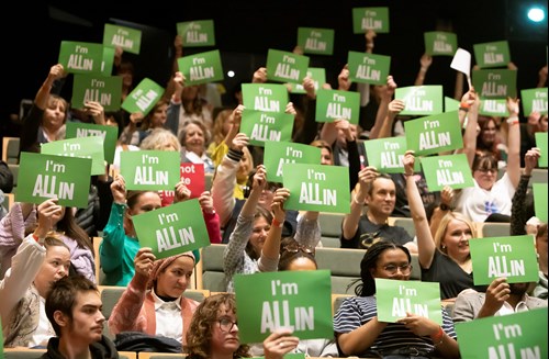 A crowd sat in an auditorium holding up green A4 sized card that says "I'm All In" in white writing.