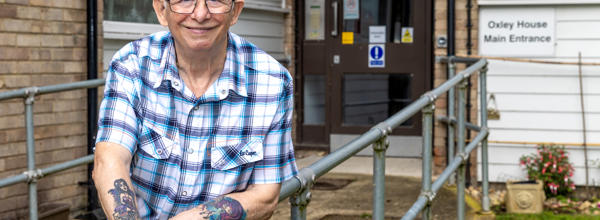 An older Eastlight resident stands outside one of Eastlight’s Housing for Older People schemes, leaning on a metal rail, smiling at the camera. 