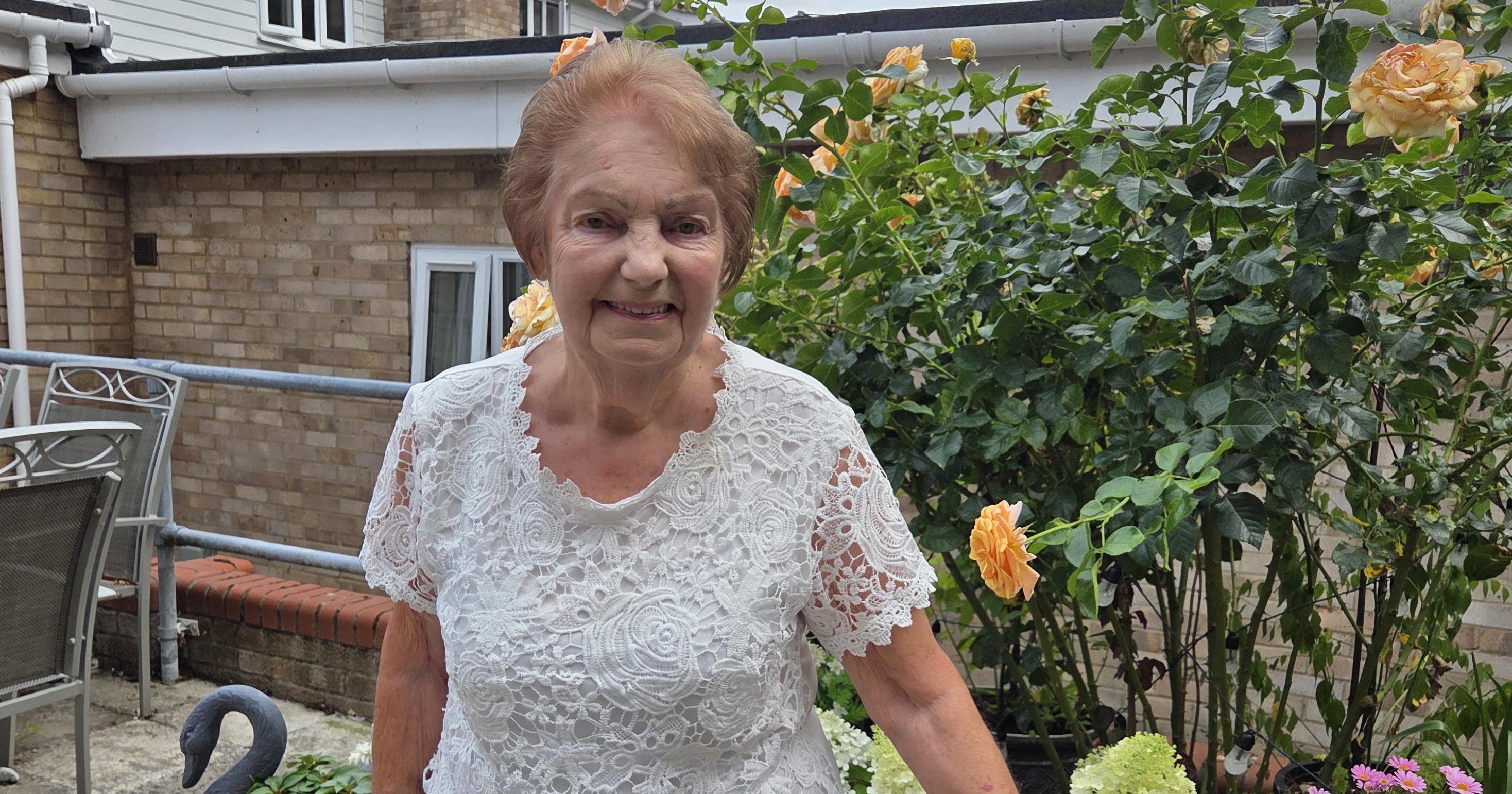 An older lady is standing in front of a flower bed. She is holding a walking stick.