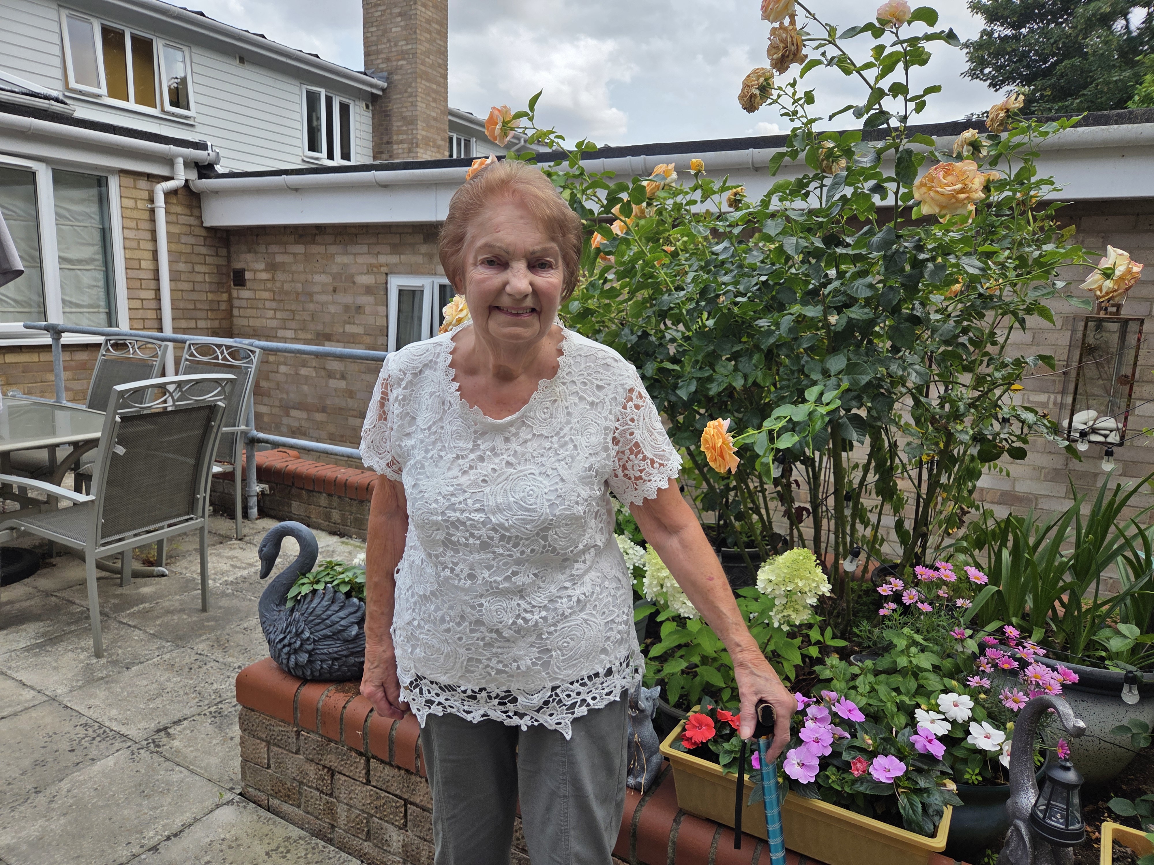 An older lady is standing in front of a flower bed. She is holding a walking stick.