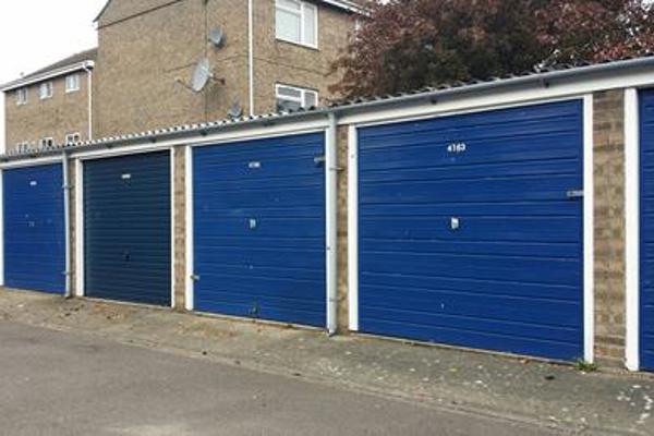  A row of blue garages, now as 'Dewlands Garages'. In front of them is some grey tarmac, and in the background, some houses. 