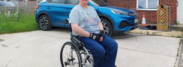 Eastlight resident, Craig, sitting in his wheelchair on his driveway, smiling at the camera. In the background is Craig's house and a blue car. Craig is wearing a grey t-shirt, blue jeans and black glasses.