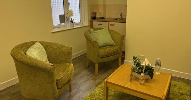A cosy room with two small armchairs around a small coffee table. A sink and cupboards are situated behind the chairs.