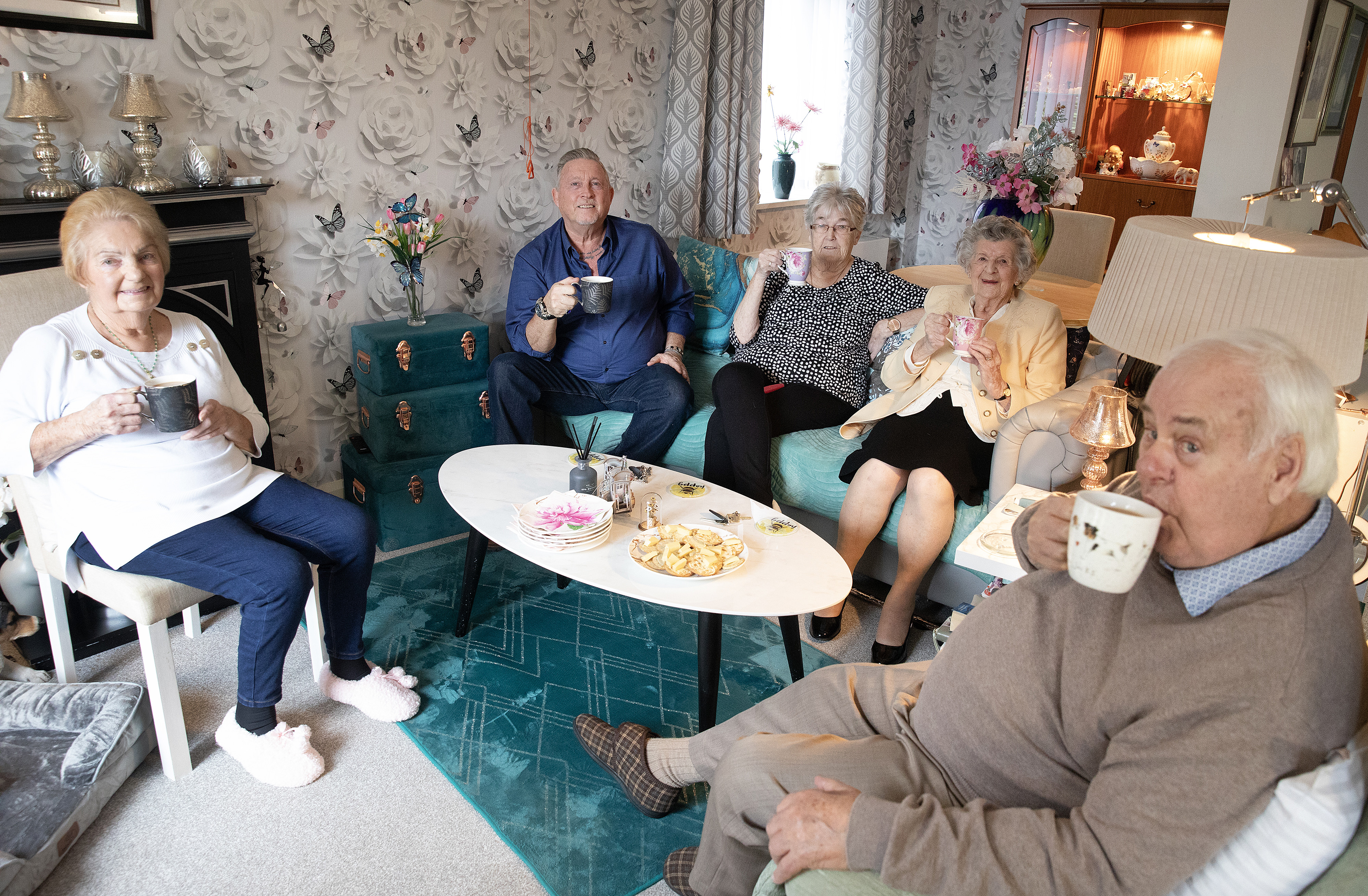 A group of older residents sitting together in a sitting room drinking tea. There are cakes on the table in the middle and various ornaments and pieces of furniture in the background. 
