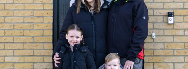 An Eastlight family, including a Mum, Dad, and two young children, standing huddled together on their front doorstep, smiling at the camera. Behind them is a smart, black front door, and, around the door, some modern-looking, light-brown brickwork. 