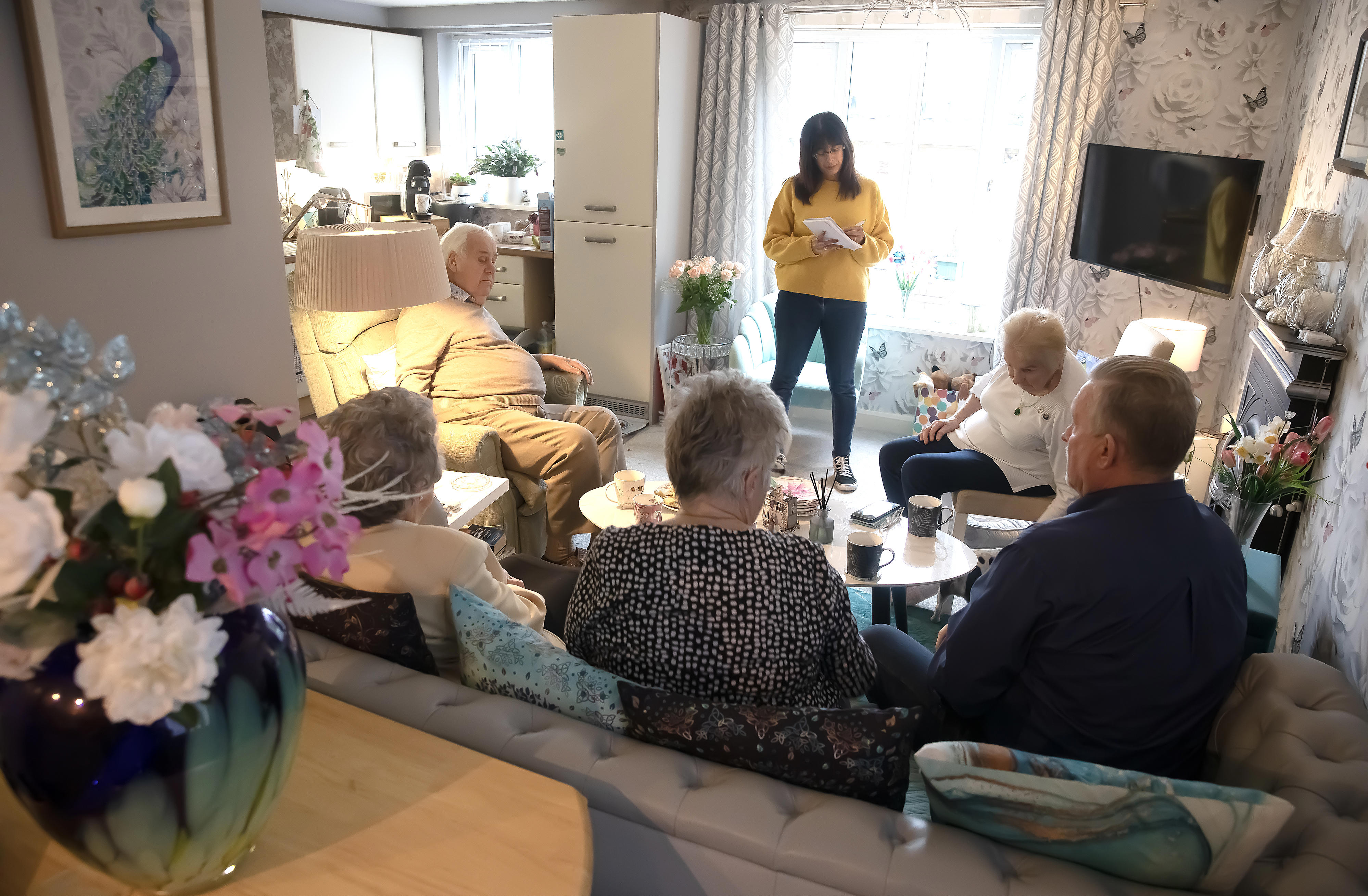 A group of older residents sitting inside a living room within one of our housing for older people schemes. The photo is taken from behind, so you cannot see their faces. They are looking towards an Eastlight staff member, who is standing in front of them, wearing a yellow jumper and dark trousers. She is looking down, taking notes on a notepad. 