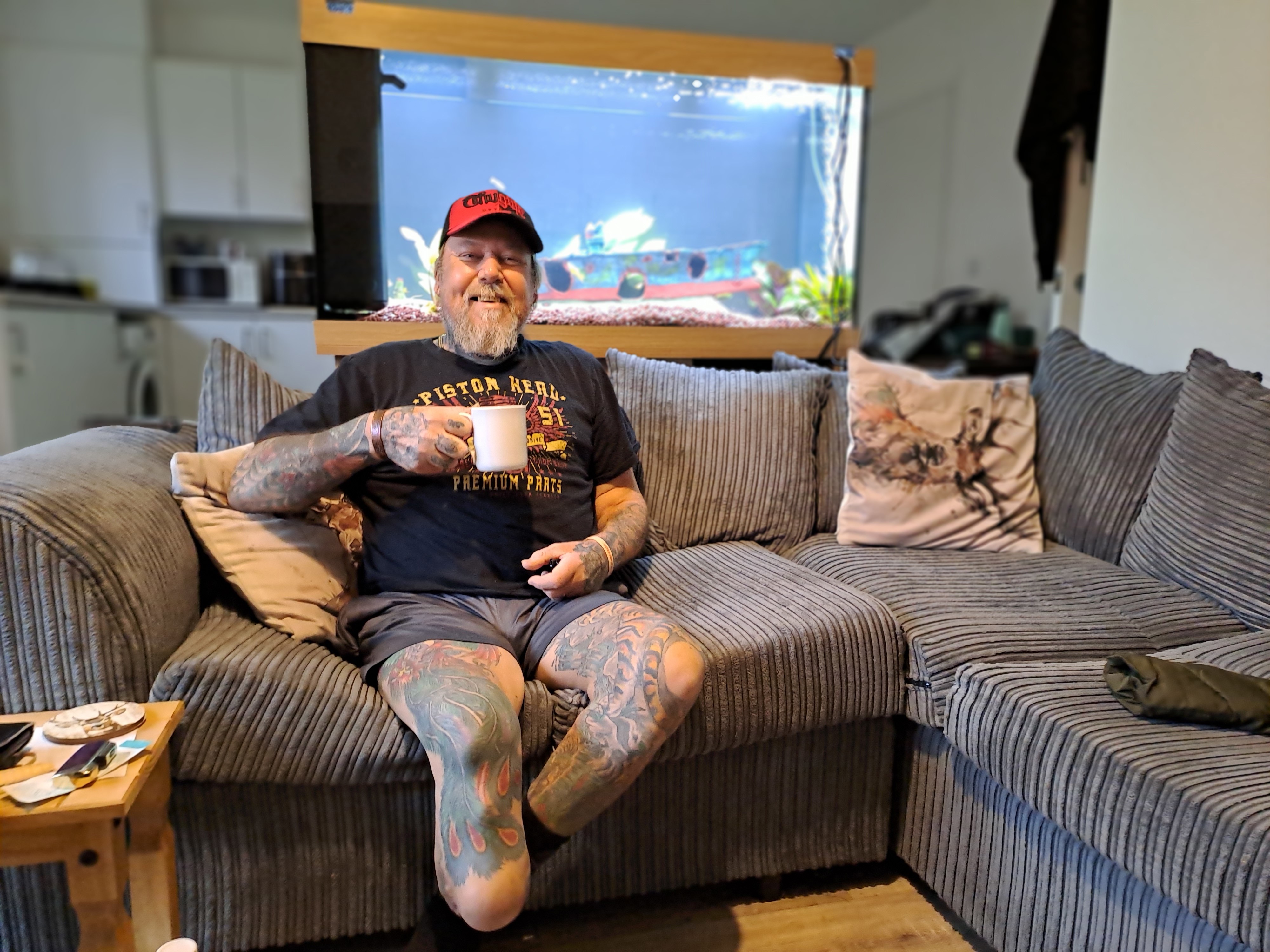 A man with a beard and tattoos over his arms and legs is sat on a grey sofa holding up a white mug and smiling.